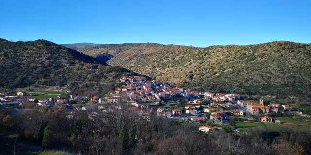 Vue d’ensemble d’un village aux toits ocres installé sur une pente, entouré de collines sèches et d’un ciel bleu dégagé.