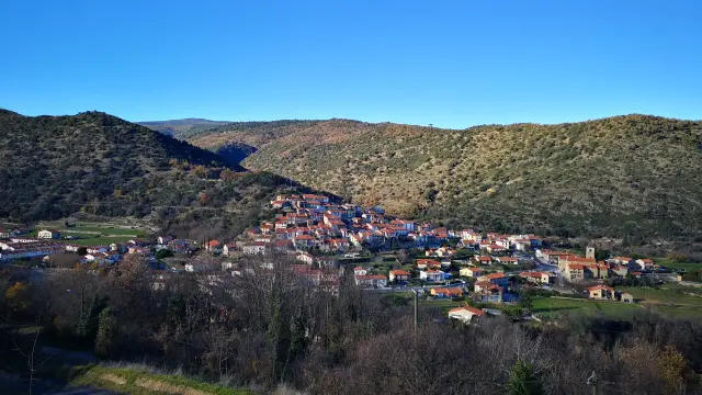 Vue d’ensemble d’un village aux toits ocres installé sur une pente, entouré de collines sèches et d’un ciel bleu dégagé.