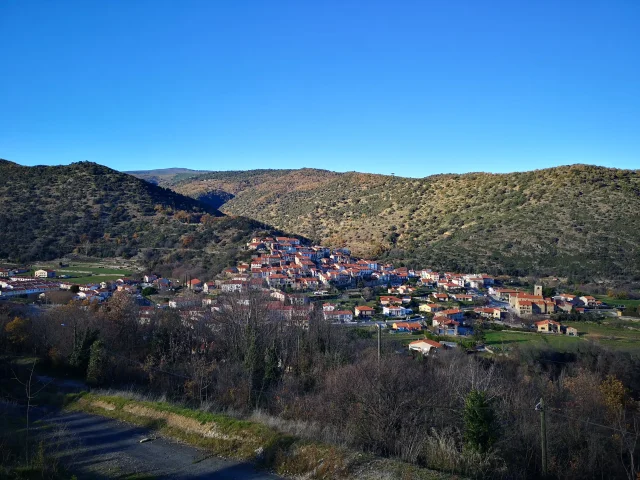 Vue d’ensemble d’un village aux toits ocres installé sur une pente, entouré de collines sèches et d’un ciel bleu dégagé.