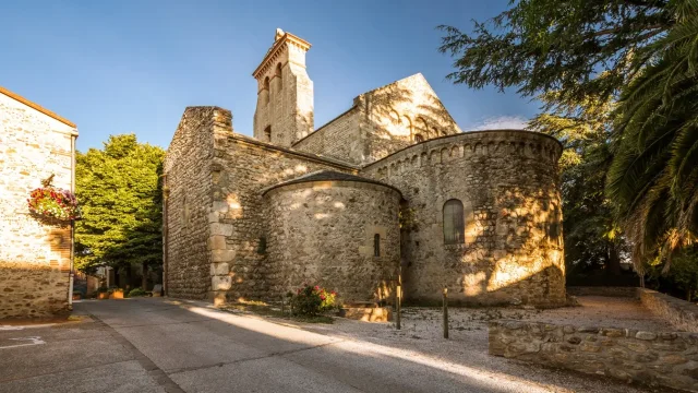 Église romane en pierre, éclairée par le soleil, avec abside arrondie et clocher, dans une rue calme bordée d’arbres.
