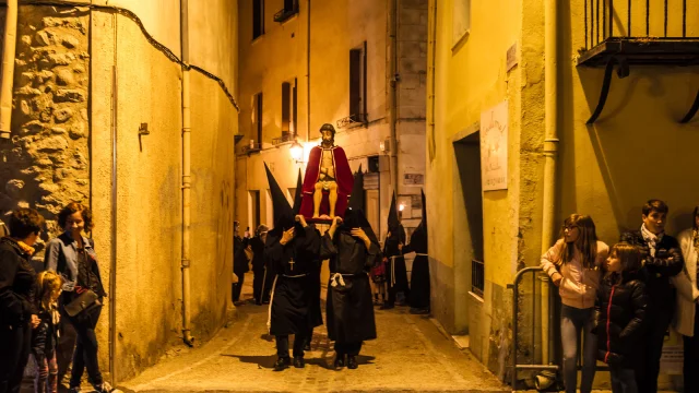 Pénitents en noir portant une statue du Christ assis et couronné d’épines dans une ruelle étroite, observés par des habitants.