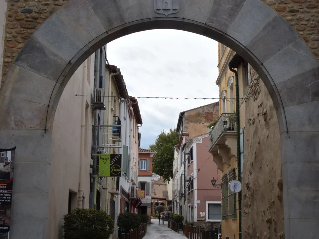 Rue pavée vue à travers une grande arche en pierre, bordée de maisons colorées et de commerces.