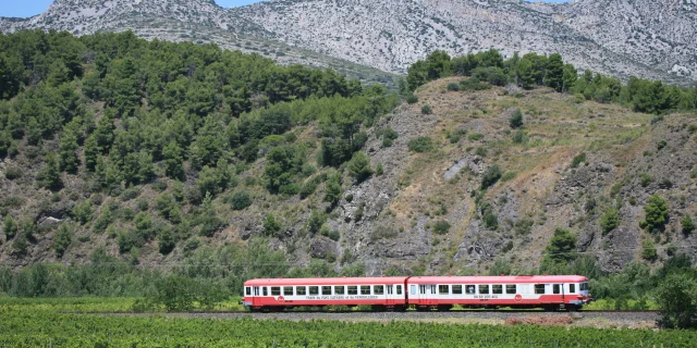 Train rouge traversant les vignes au pied des collines rocailleuses.