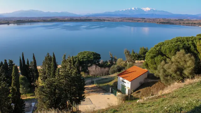 Vue d’un grand lac calme bordé d’arbres, avec une petite maison au toit orange au premier plan et des montagnes enneigées à l’horizon.