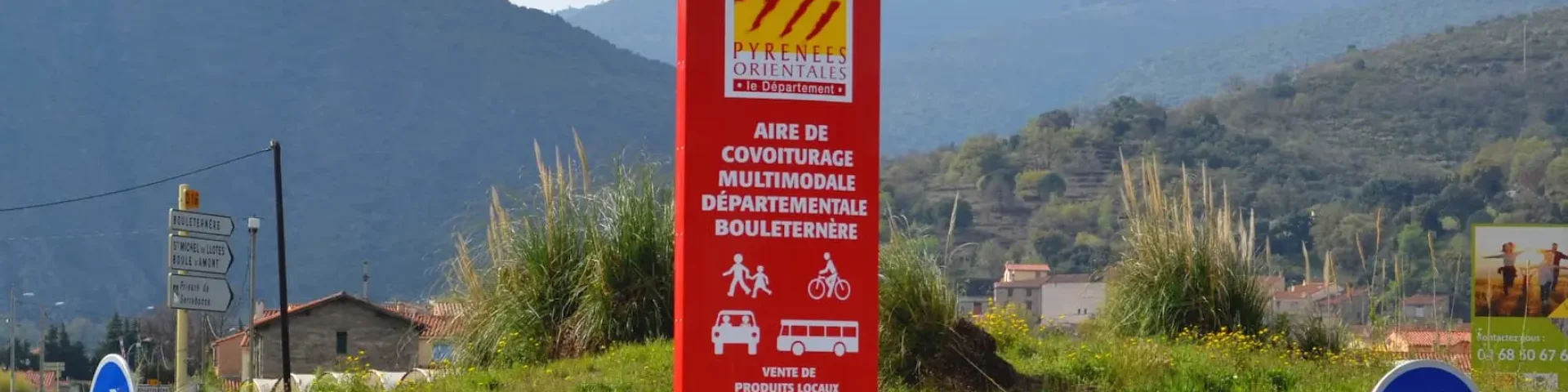 Panneau rouge indiquant l’aire de covoiturage multimodale départementale de Bouleternère, avec les pictogrammes de marcheurs, cyclistes, voiture et bus, sur fond de montagnes verdoyantes des Pyrénées-Orientales.