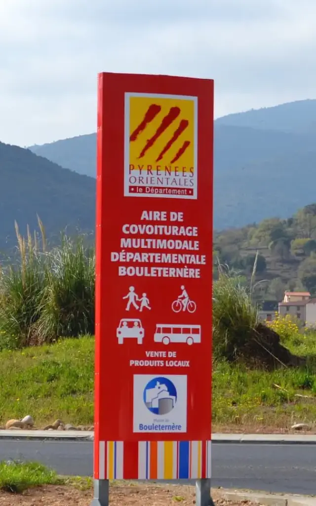 Panneau rouge indiquant l’aire de covoiturage multimodale départementale de Bouleternère, avec les pictogrammes de marcheurs, cyclistes, voiture et bus, sur fond de montagnes verdoyantes des Pyrénées-Orientales.