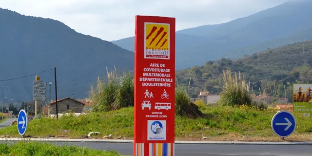 Panneau rouge indiquant l’aire de covoiturage multimodale départementale de Bouleternère, avec les pictogrammes de marcheurs, cyclistes, voiture et bus, sur fond de montagnes verdoyantes des Pyrénées-Orientales.