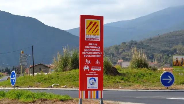 Panneau rouge indiquant l’aire de covoiturage multimodale départementale de Bouleternère, avec les pictogrammes de marcheurs, cyclistes, voiture et bus, sur fond de montagnes verdoyantes des Pyrénées-Orientales.