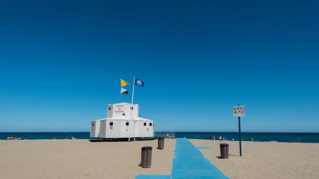 Poste de secours sur une plage avec chemin d’accès bleu menant vers la mer sous un ciel dégagé.