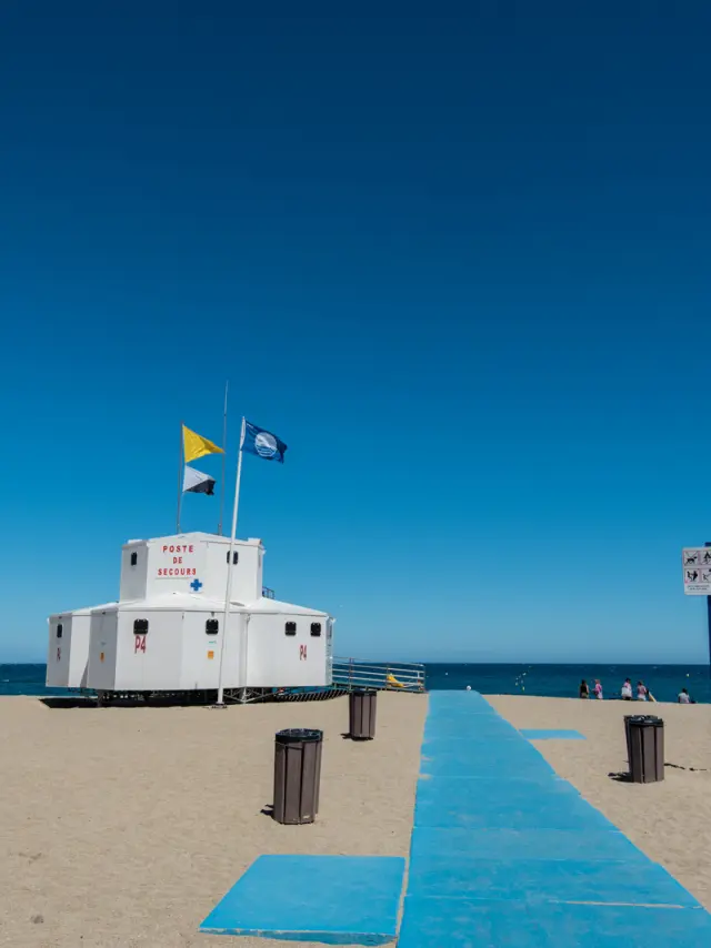Poste de secours sur une plage avec chemin d’accès bleu menant vers la mer sous un ciel dégagé.