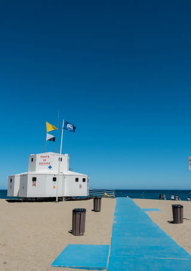 Poste de secours sur une plage avec chemin d’accès bleu menant vers la mer sous un ciel dégagé.