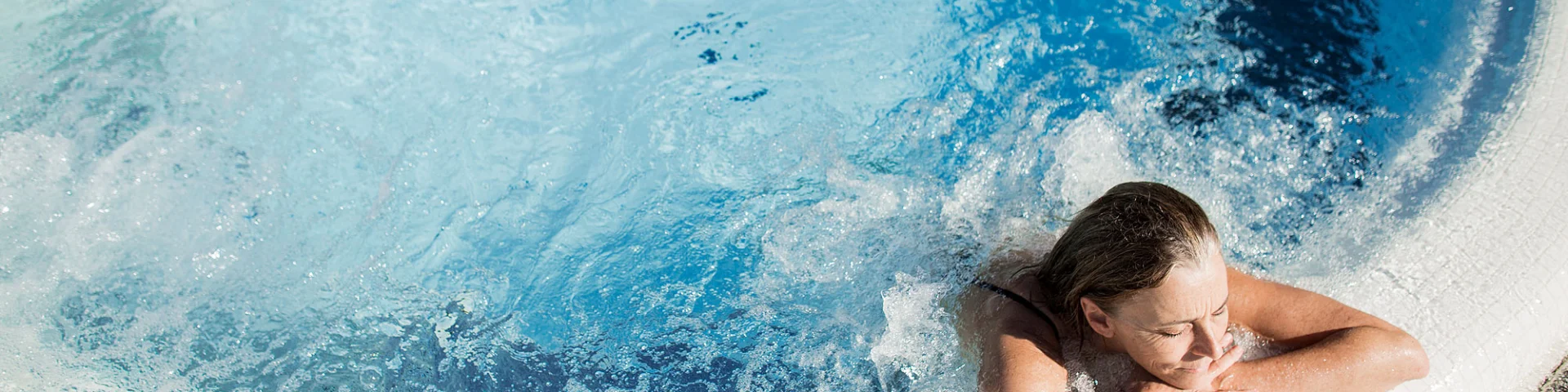 Femme se relaxant dans un bassin à remous en extérieur, appuyée sur le bord du jacuzzi, sous un soleil éclatant.