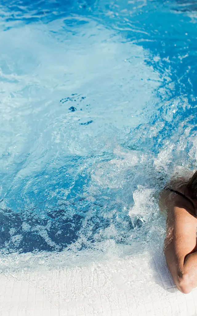 Femme se relaxant dans un bassin à remous en extérieur, appuyée sur le bord du jacuzzi, sous un soleil éclatant.