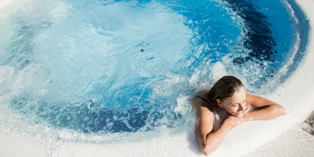 Femme se relaxant dans un bassin à remous en extérieur, appuyée sur le bord du jacuzzi, sous un soleil éclatant.