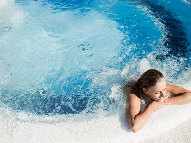 Femme se relaxant dans un bassin à remous en extérieur, appuyée sur le bord du jacuzzi, sous un soleil éclatant.