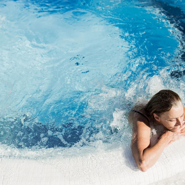 Femme se relaxant dans un bassin à remous en extérieur, appuyée sur le bord du jacuzzi, sous un soleil éclatant.