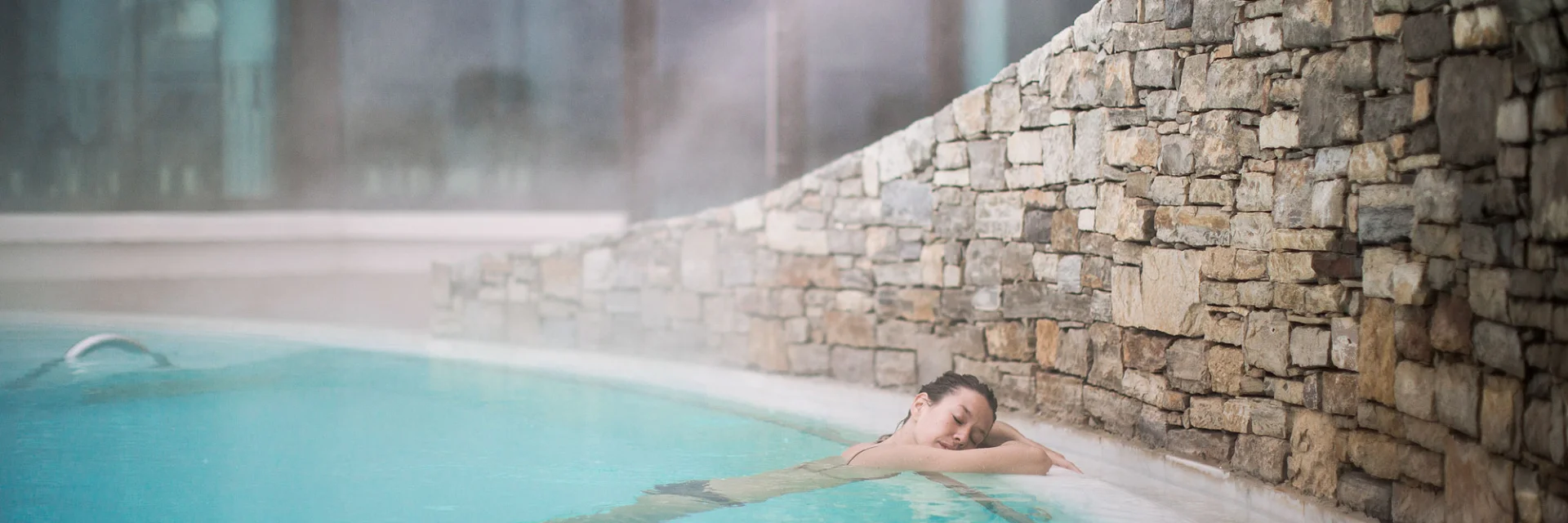 Femme se relaxant dans un bassin d’eau chaude intérieure, adossée à un mur en pierres, dans une ambiance paisible et légèrement brumeuse.