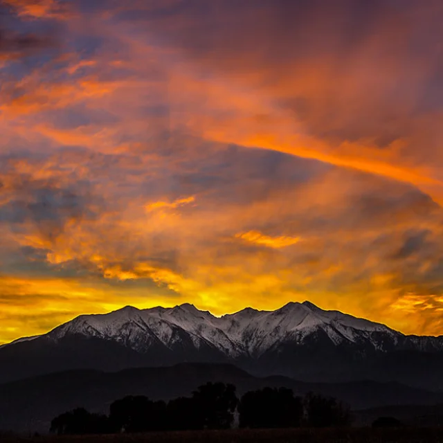 Le massif du Canigó enneigé vu depuis Ponteilla, au lever du soleil, sous un ciel flamboyant aux teintes orange, jaune et violette.