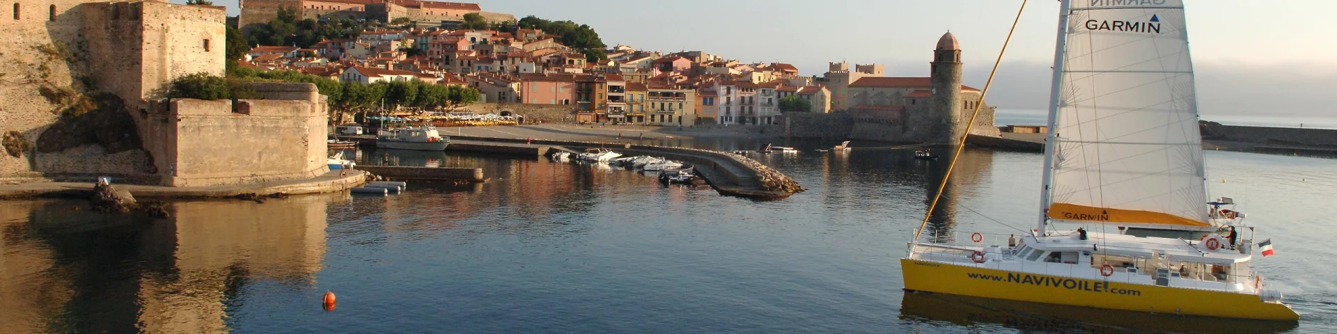 Catamaran jaune naviguant dans une baie calme près d’une ville côtière et d’un port de plaisance.