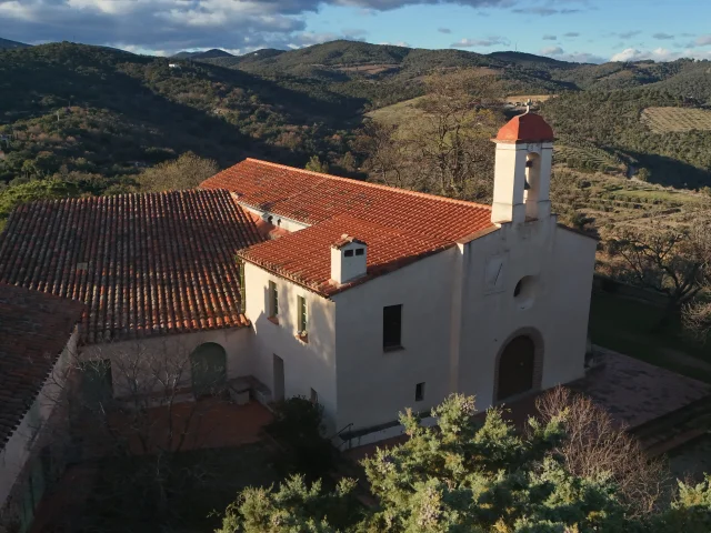 Ermitage à toit de tuiles rouges sur un versant boisé, entouré de collines et de vallons sous un ciel partiellement nuageux.