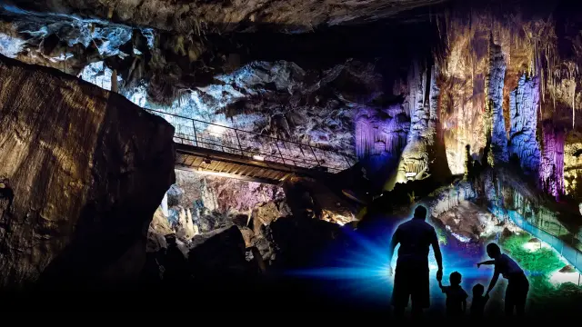 Intérieur d’une grotte éclairée de couleurs avec un pont métallique au-dessus de formations rocheuses et quatre silhouettes au premier plan.