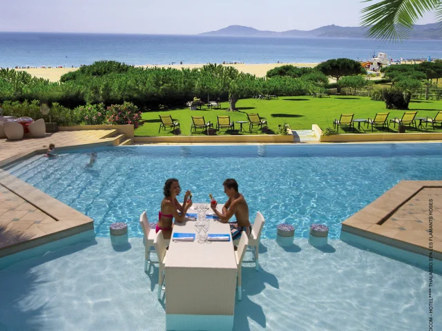 Couple attablé dans une piscine, partageant un verre face à la mer Méditerranée, avec pelouse, transats et plage en arrière-plan.