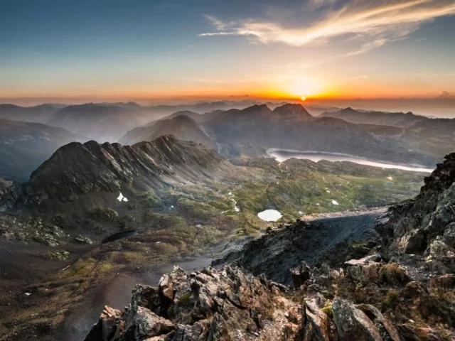 Paysage de haute montagne au lever du soleil, avec crêtes rocheuses, lacs d’altitude et vallées noyées dans la brume.