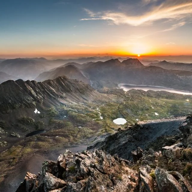 Paysage de haute montagne au lever du soleil, avec crêtes rocheuses, lacs d’altitude et vallées noyées dans la brume.