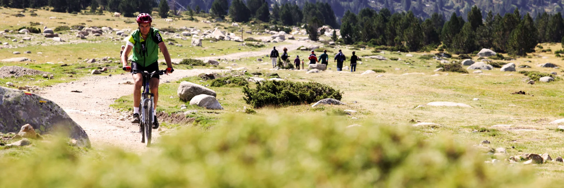 Cycliste en VTT avançant sur un sentier de montagne, avec un groupe de marcheurs au loin dans un paysage rocailleux.