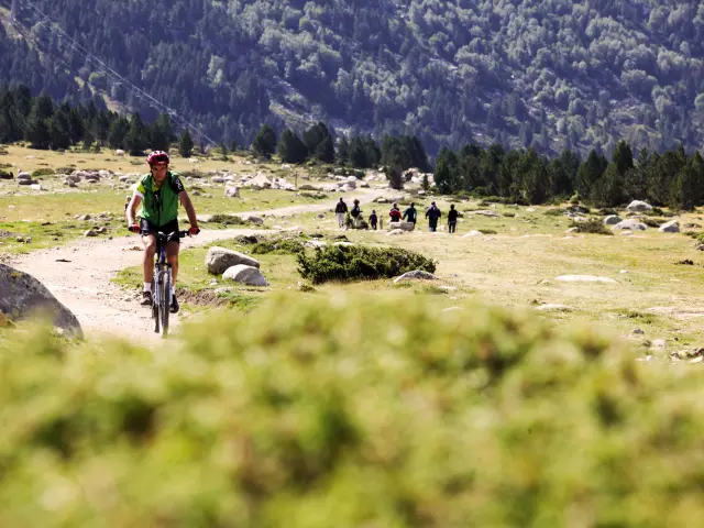 Cycliste en VTT avançant sur un sentier de montagne, avec un groupe de marcheurs au loin dans un paysage rocailleux.