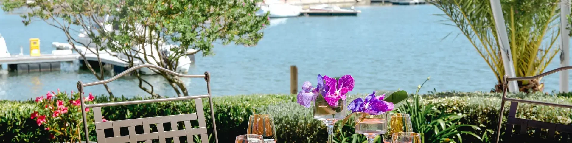 Table dressée en terrasse face à un plan d’eau bordé de palmiers et de bateaux de plaisance.