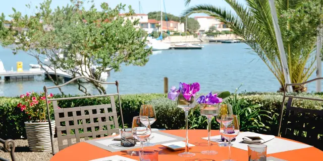 Table dressée en terrasse face à un plan d’eau bordé de palmiers et de bateaux de plaisance.
