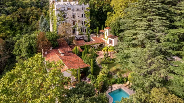 Vue aérienne d’un grand bâtiment en pierre entouré de toits en tuiles, d’un jardin arboré et d’une piscine, au milieu d’une forêt dense.