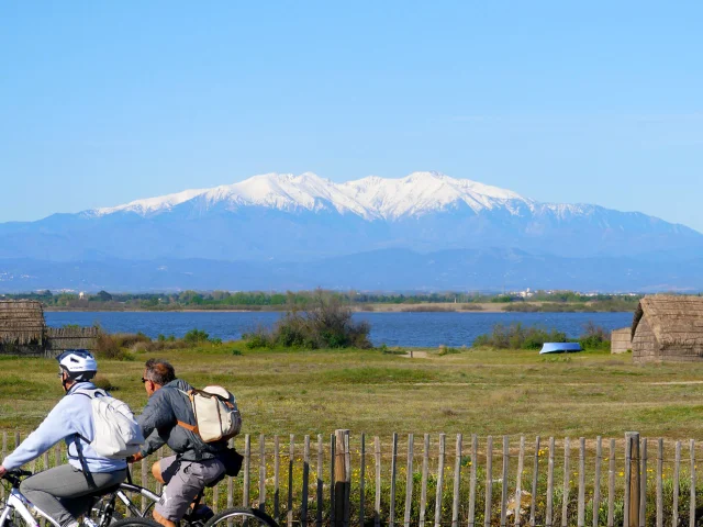 Deux cyclistes roulent près d’un étang, avec cabanes en roseaux et montagnes enneigées au loin.
