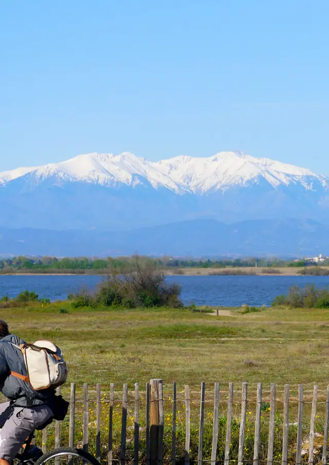 Deux cyclistes roulent près d’un étang, avec cabanes en roseaux et montagnes enneigées au loin.