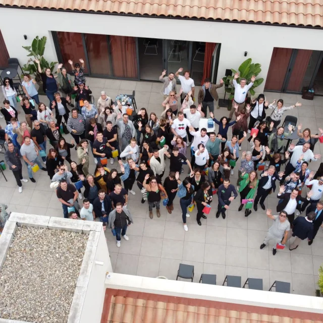 Photo aérienne d’un grand groupe de participants réunis dans la cour intérieure du Camping La Sirène à Argelès-sur-Mer lors d’un séminaire professionnel, levant la main vers l’objectif.