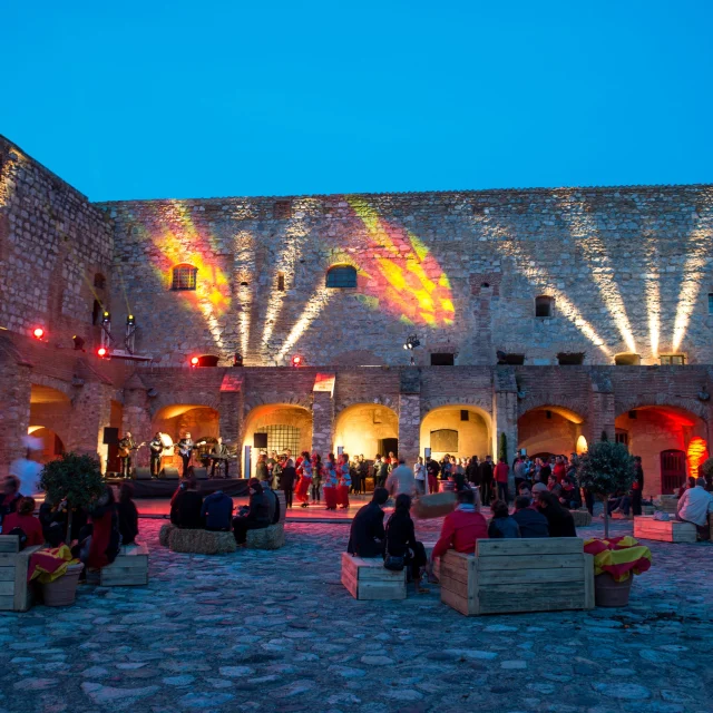 Soirée animée dans la cour intérieure de la forteresse de Salses, éclairée par des projections lumineuses rouges et jaunes sur les murs de pierre, avec des spectateurs assis et un groupe sur scène.