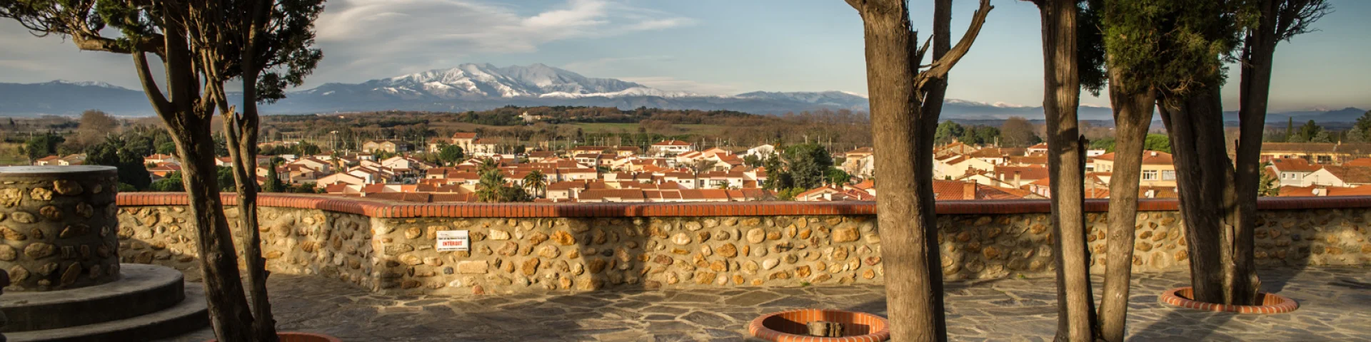 Terrasse en pierre bordée d’arbres offrant une vue sur le village et les montagnes enneigées au loin.