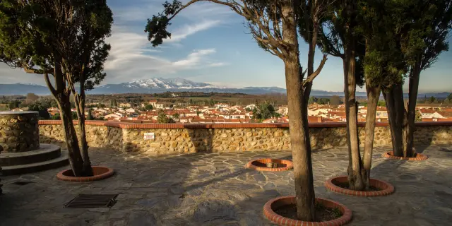 Terrasse en pierre bordée d’arbres offrant une vue sur le village et les montagnes enneigées au loin.