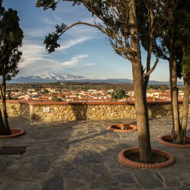 Terrasse en pierre bordée d’arbres offrant une vue sur le village et les montagnes enneigées au loin.