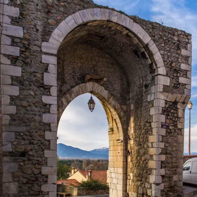 Porte médiévale en pierre formant une arche ouvrant sur les toits du village et les montagnes lointaines.