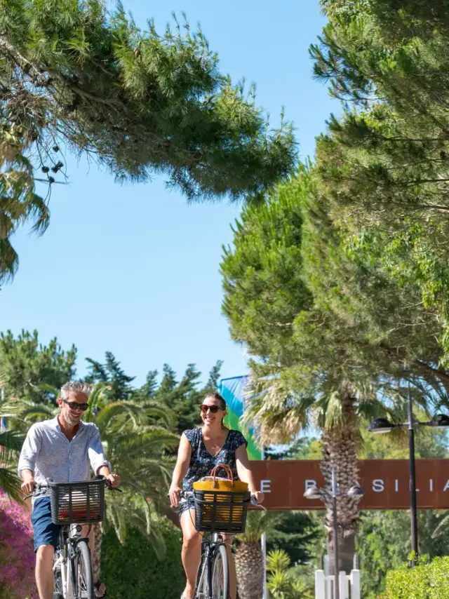 Couple à vélo circulant sur une allée bordée d’arbres et de végétation au camping Le Brasilia.