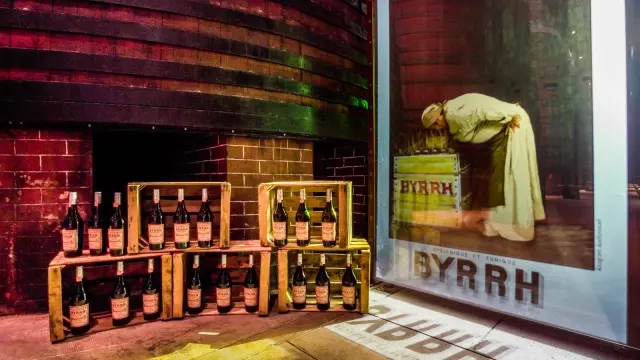 Caisse de bouteilles Byrrh disposées devant une grande affiche publicitaire ancienne dans la cave de Thuir.