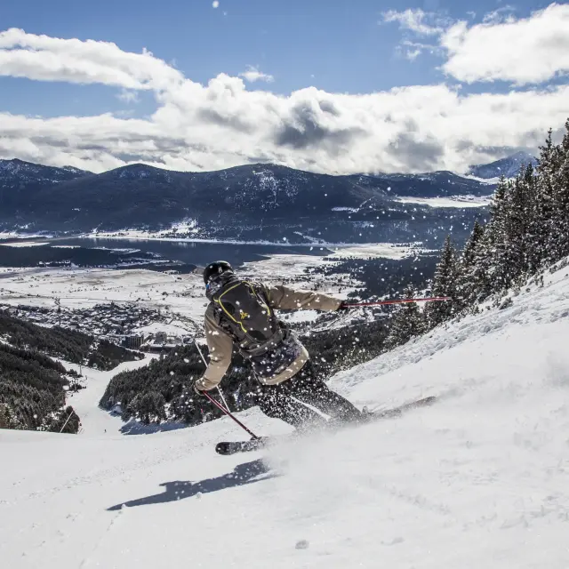 Skieur en descente sur une grande piste au-dessus du village enneigé des Angles et du lac de Matemale.