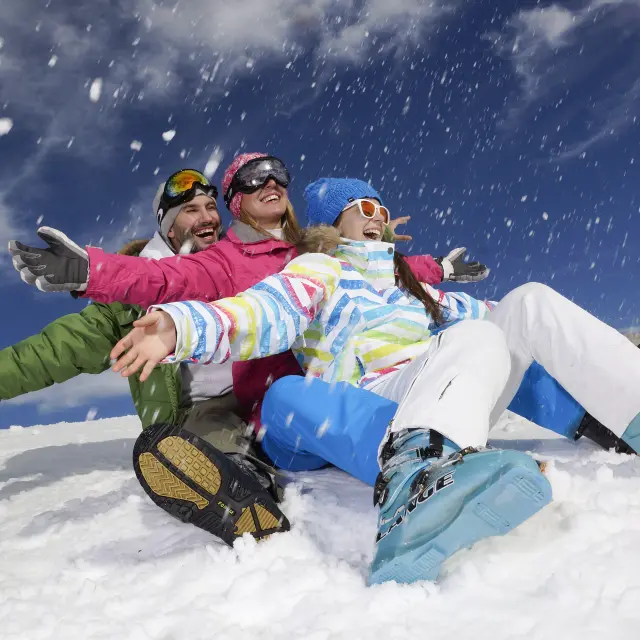 Trois adultes jouent dans la neige en combinaison de ski, bras écartés, sous un ciel bleu avec des projections de neige.