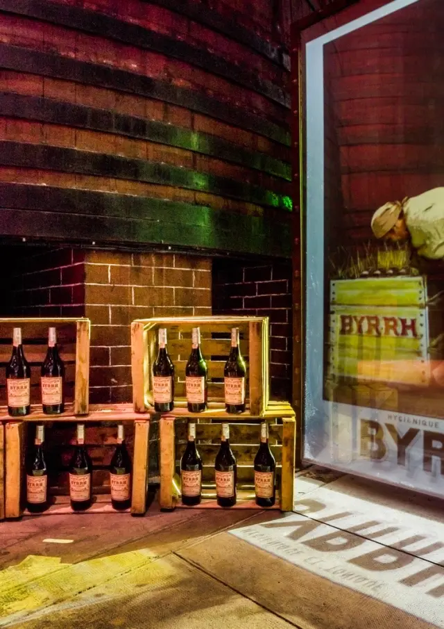 Caisse de bouteilles Byrrh disposées devant une grande affiche publicitaire ancienne dans la cave de Thuir.