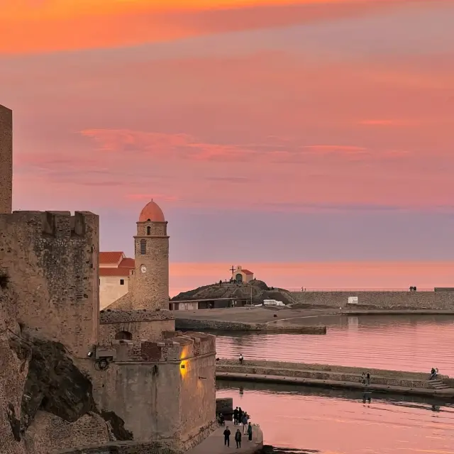 Vue de Collioure au coucher du soleil, avec les remparts du château, l'église Notre-Dame-des-Anges et le port, sous un ciel aux couleurs chaudes.