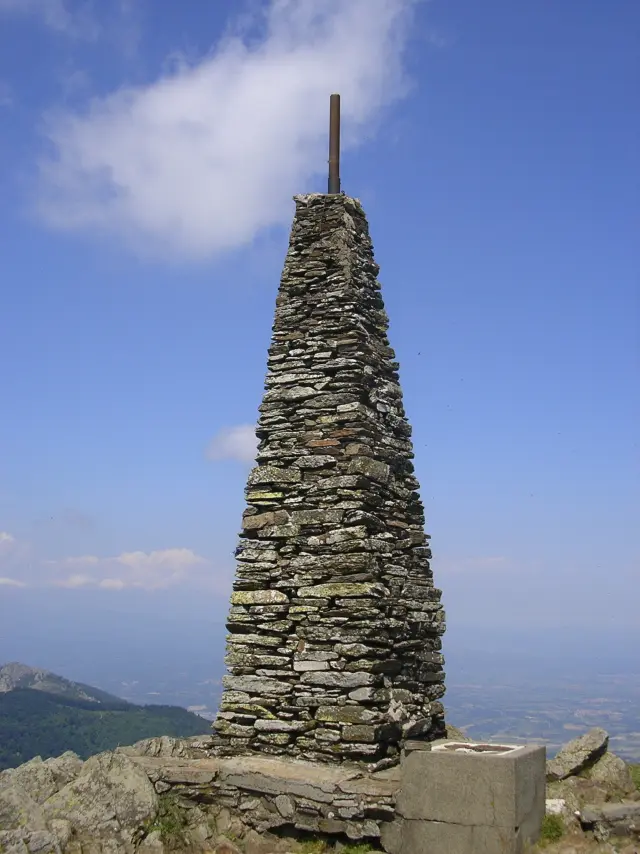 Cairn pyramidal en pierres sèches surmonté d'un mât métallique au sommet du Puig Neulós, sous un ciel bleu avec quelques nuages.