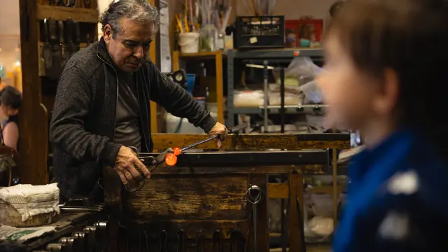 Maître verrier travaillant une masse de verre en fusion incandescente à l'aide de pinces, dans son atelier, sous le regard attentif d'un enfant.