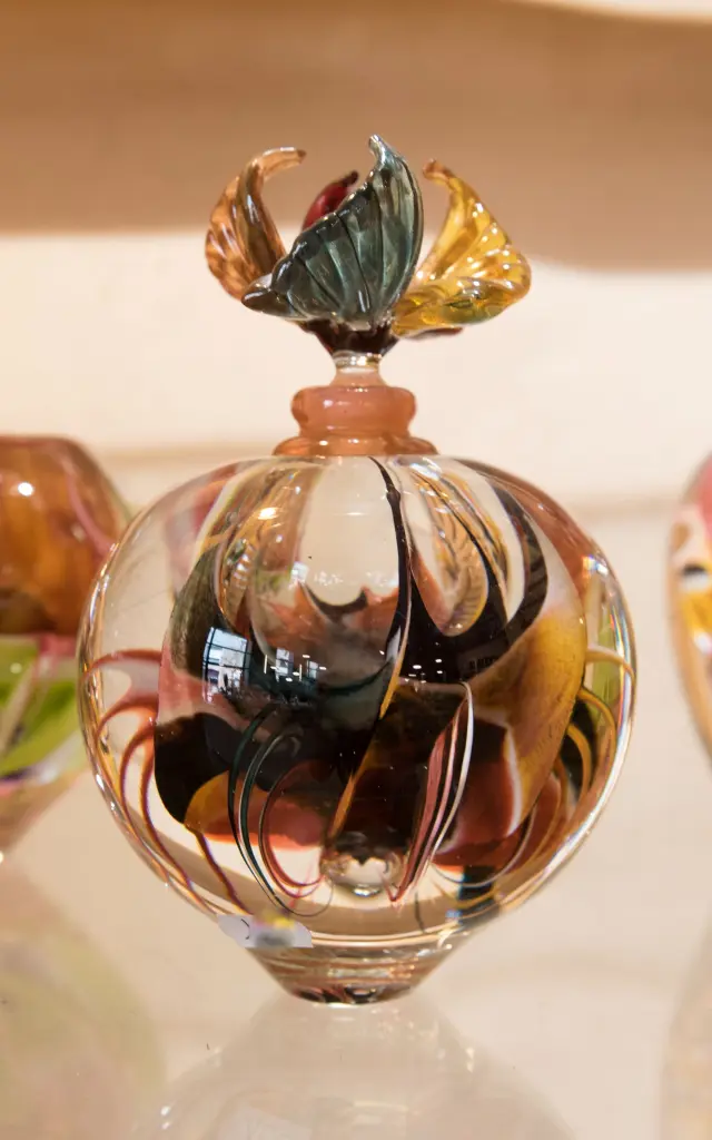Trois pièces en verre soufflé aux motifs tourbillonnants multicolores exposées sur une étagère : un vase, un flacon sphérique à bouchon sculpté en forme de fleur, et un flacon ovoïde.